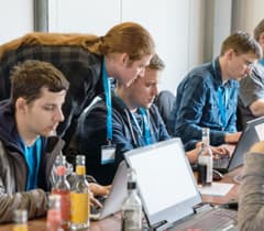 A group of developers is working together at a table, focused on their laptops. One person is leaning over to assist another developer.