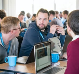 A group of developers seated at a table are engaged in a conversation, with laptops in front of them.