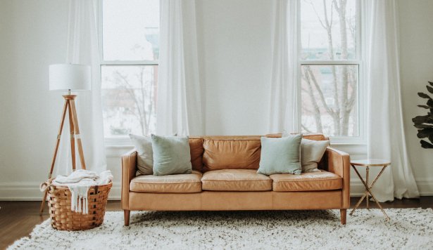 A living room features a tan leather sofa with light-colored pillows, positioned between two large windows.