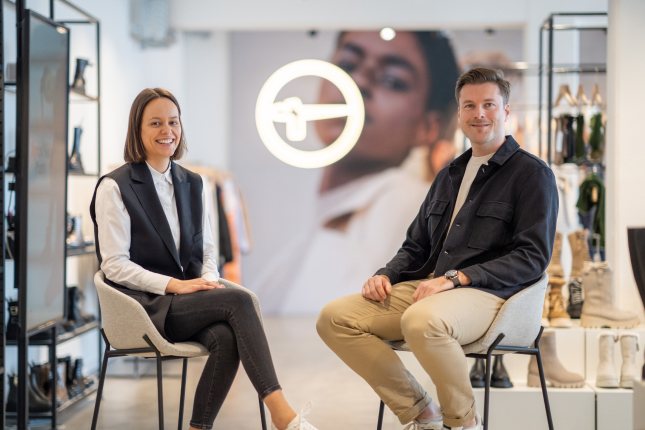 A man and a woman are sitting on chairs in a modern retail store. Behind them is the Tamaris logo. Shelves with various products are visible in the background.
