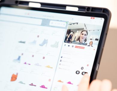 A person browses shoes on a tablet, with product images on the left and a video call on the right. The person’s finger points at an option on the screen.
